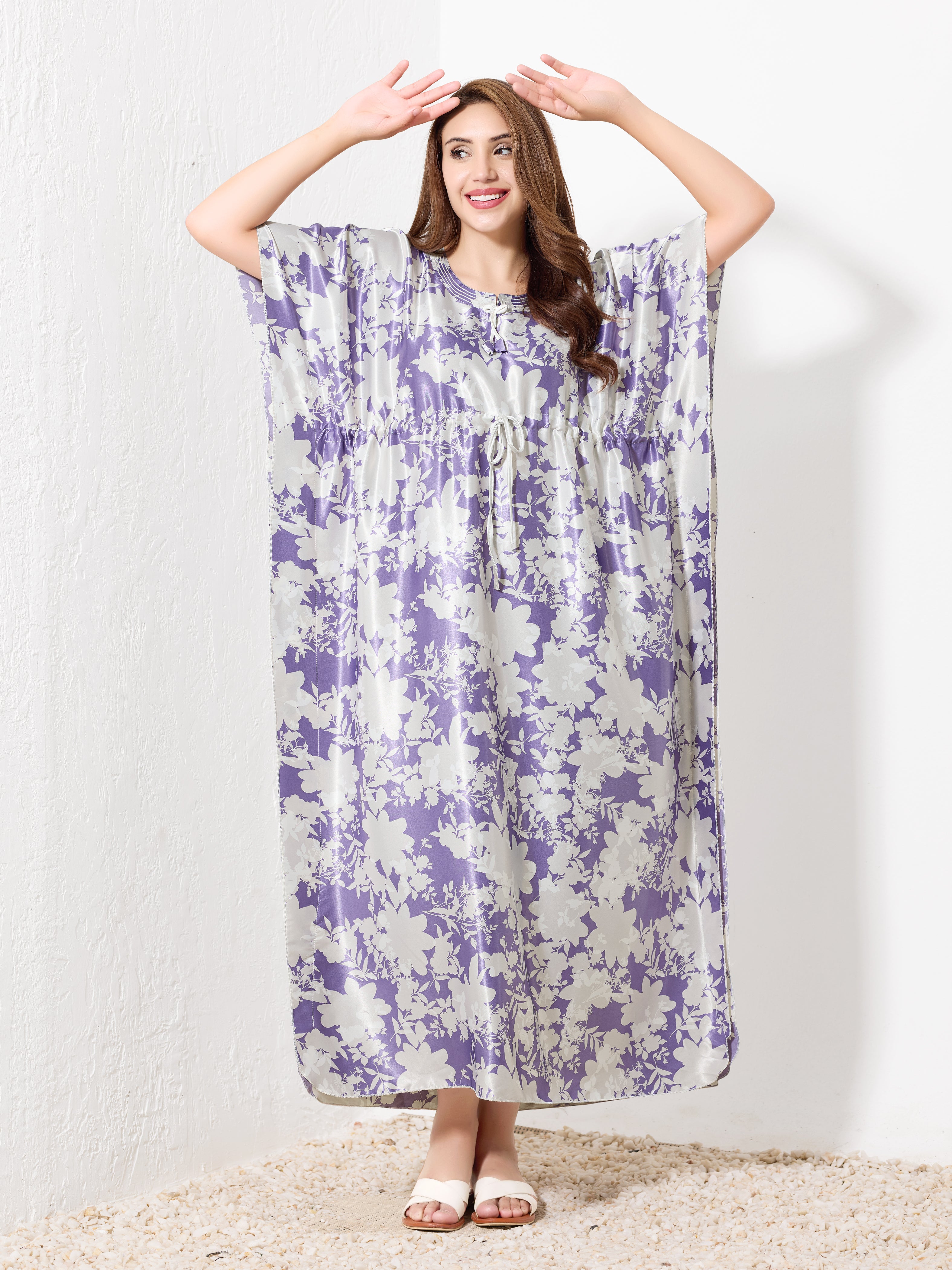 A smiling woman wears the Priincezz Nightwear Purple & White Floral Kaftan, standing against a white wall with her hands raised above her head.