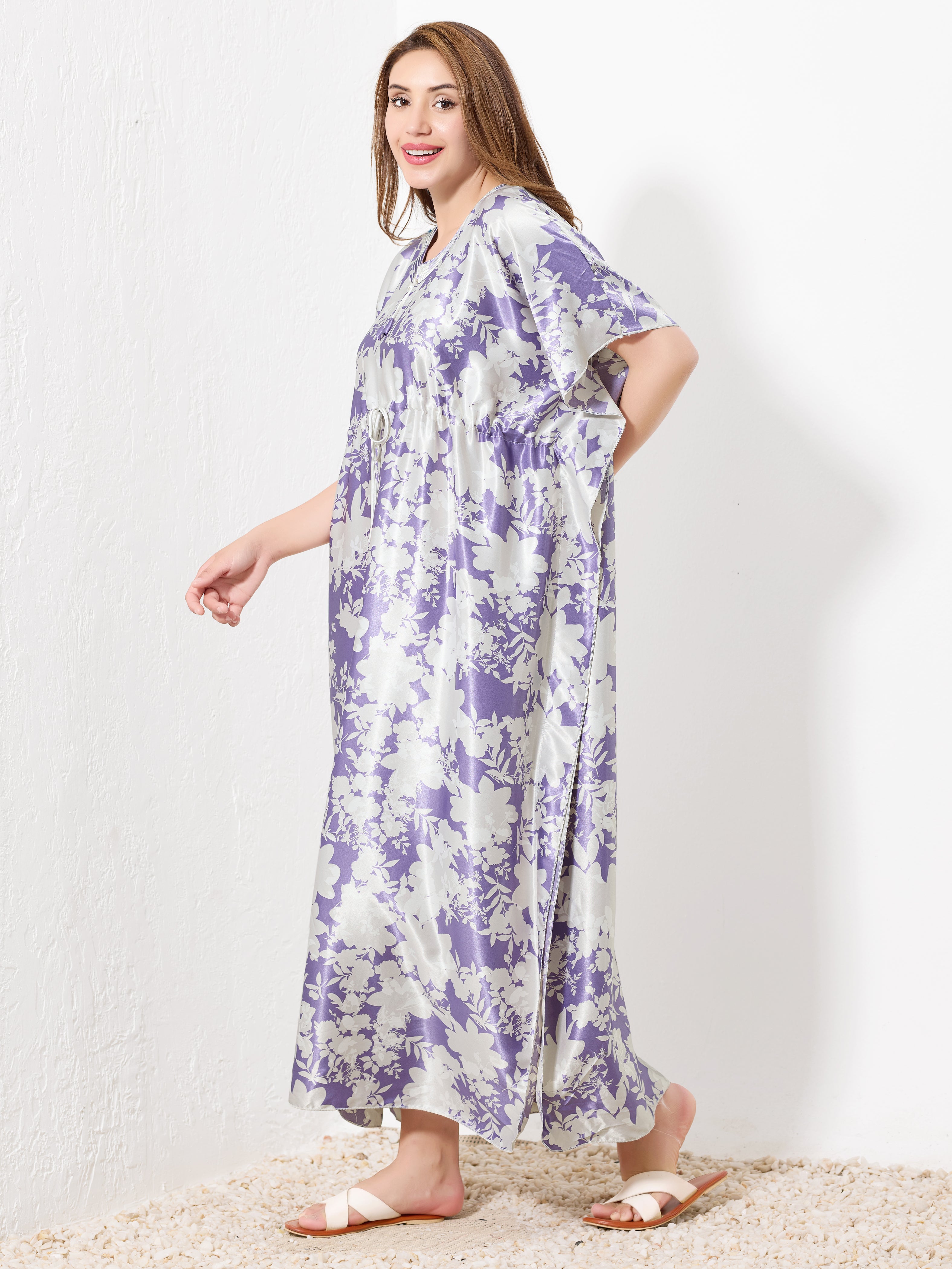 A woman models the Priincezz Nightwear Purple & White Floral Kaftan, standing barefoot on a textured floor and smiling over her shoulder—a stylish fusion of elegant loungewear and comfy nightwear.