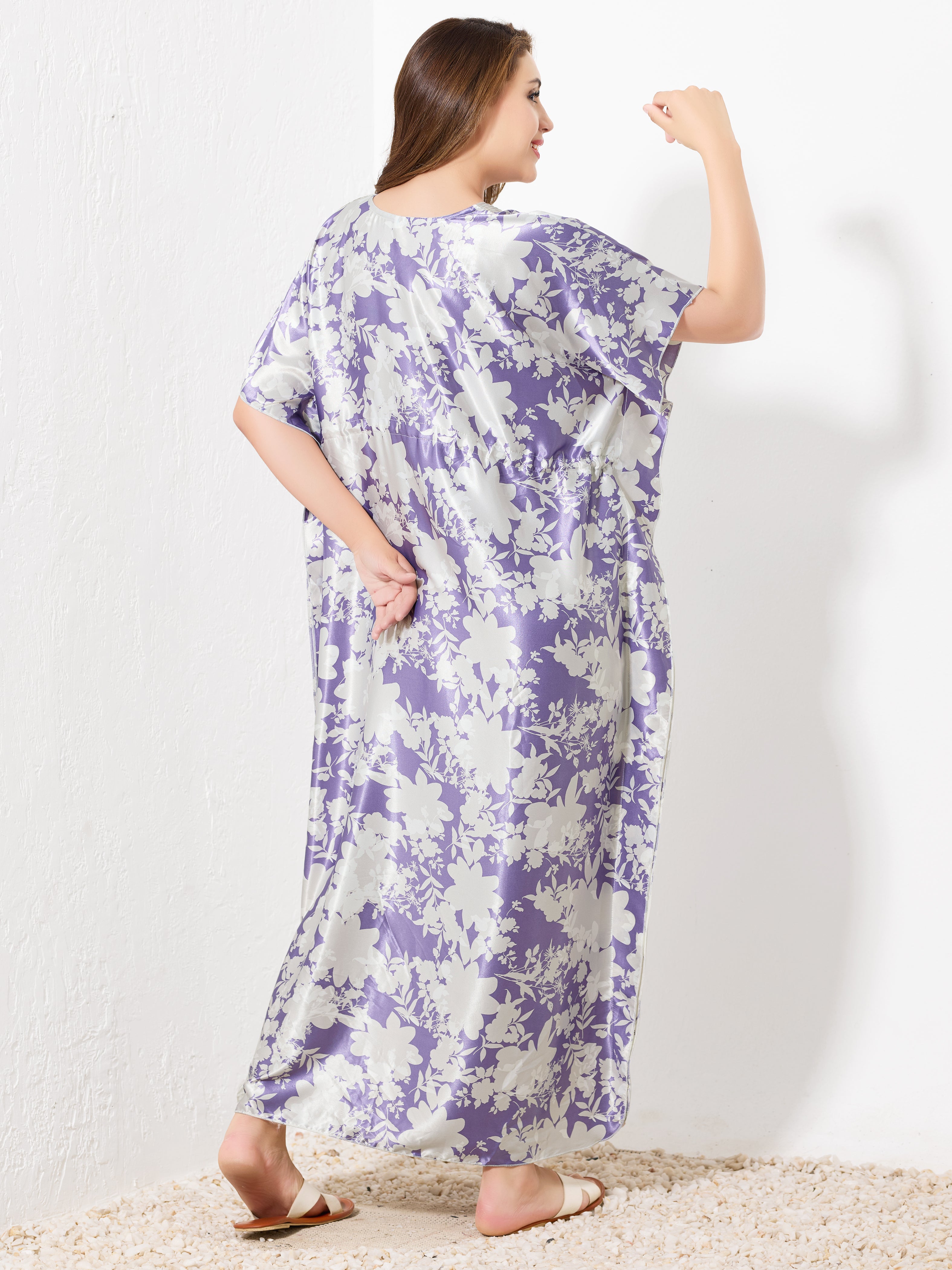 A woman stands barefoot on a textured surface, facing away. She wears the Priincezz Nightwear Purple & White Floral Kaftan—loose, floor-length satin with short sleeves—ideal for elegant and comfortable evenings. Her hair is down, one arm bent upward.